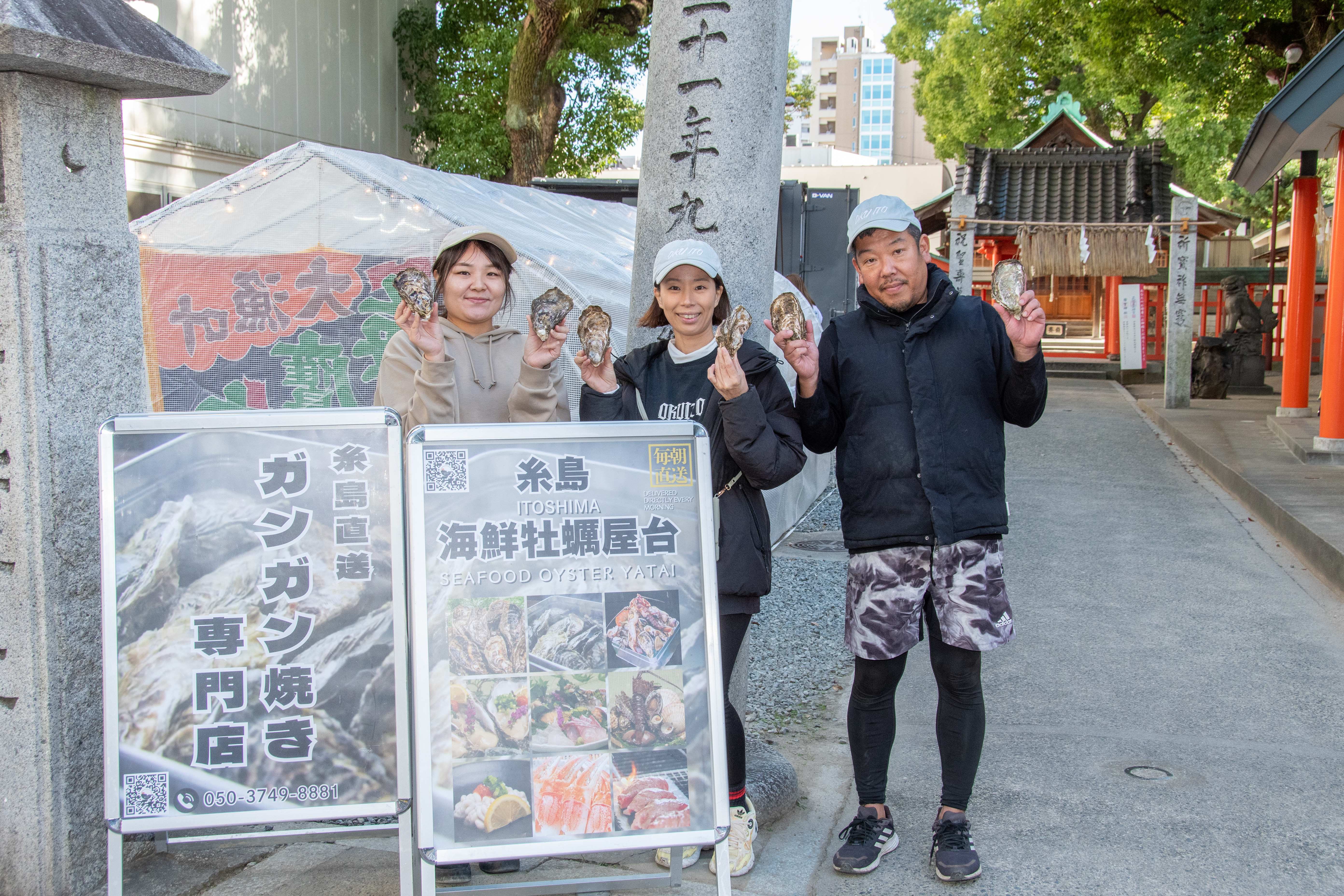 糸島直送ガンガン焼き専門店の看板の前に立つ奥糸のスタッフ3人