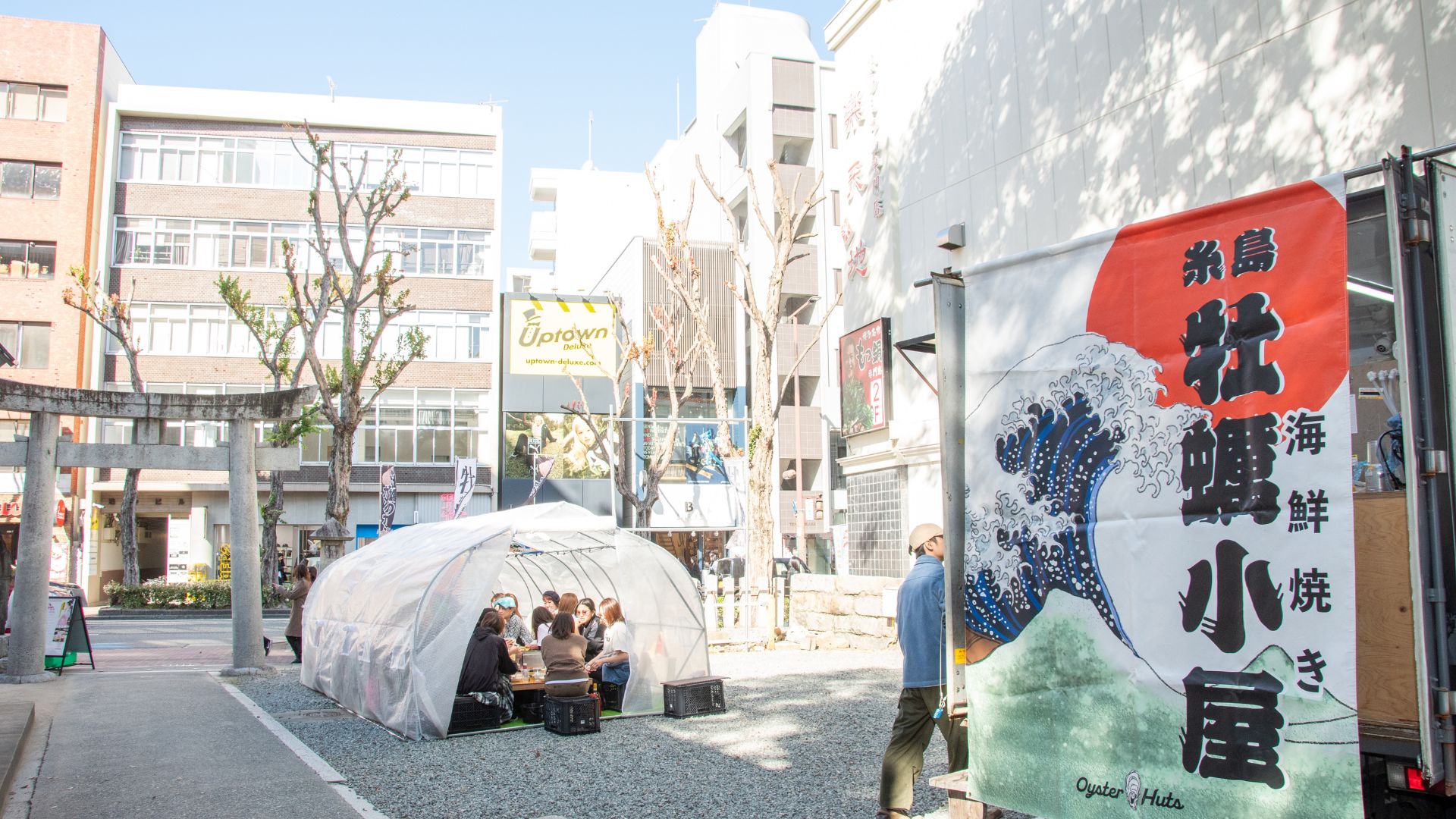都会のビルに囲まれた若宮神社の境内に、牡蠣小屋のテントと大きな「糸島牡蠣小屋」の看板が並ぶ様子。鳥居や参道が写り、神社の中で牡蠣小屋を楽しめる特別なロケーションを表現しています。
